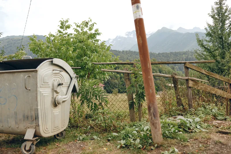 A weathered mailbox on a wheeled stand rests beside a wooden fence on a hillside, with lush vegetation and distant mountains under a calm sky.