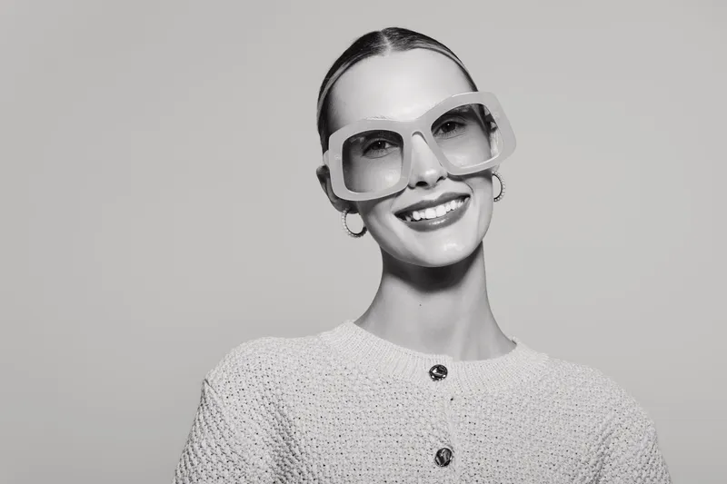 Smiling white woman with light skin wearing large sunglasses and knit sweater posing on grey background.