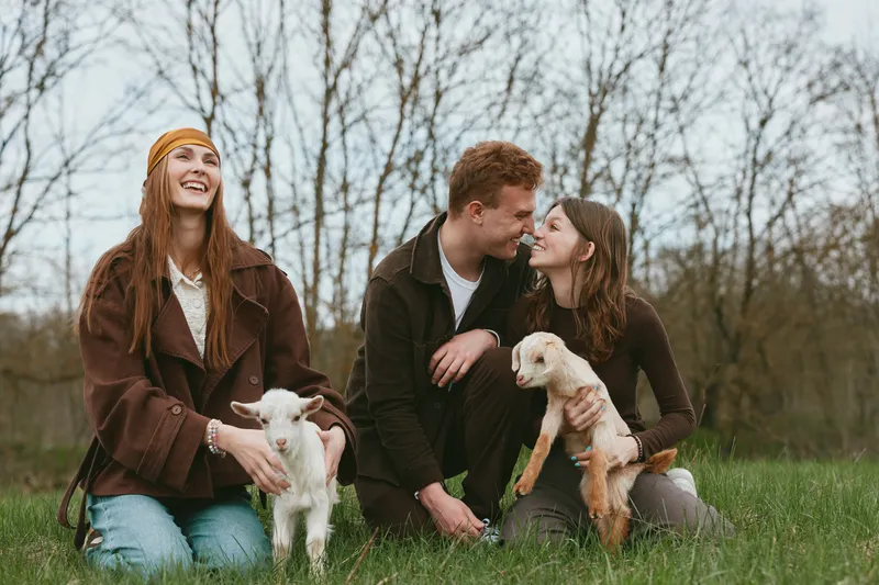 Cheerful outdoor scene with a group of friends and family, two goat kids nearby, warm casual clothing, candid laughter, natural light, grassy field, relaxed lifestyle moment