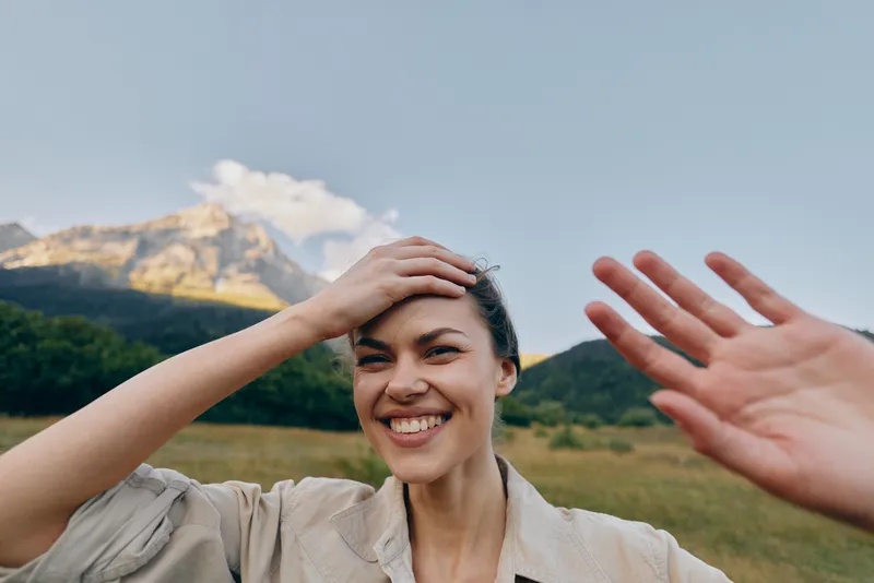 A smiling young woman shields her forehead with one hand while waving with the other in a sunny outdoor field, mountains visible behind her for a candid travel vibe.