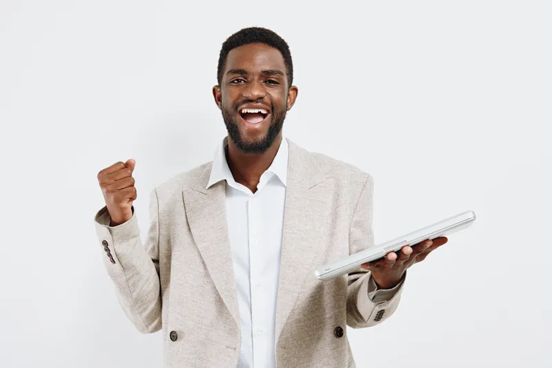 Smiling man with short curly black hair and beard, wearing a light beige checked blazer and white shirt, expressing excitement and success, holding a laptop in one hand, raising his fist, shot against