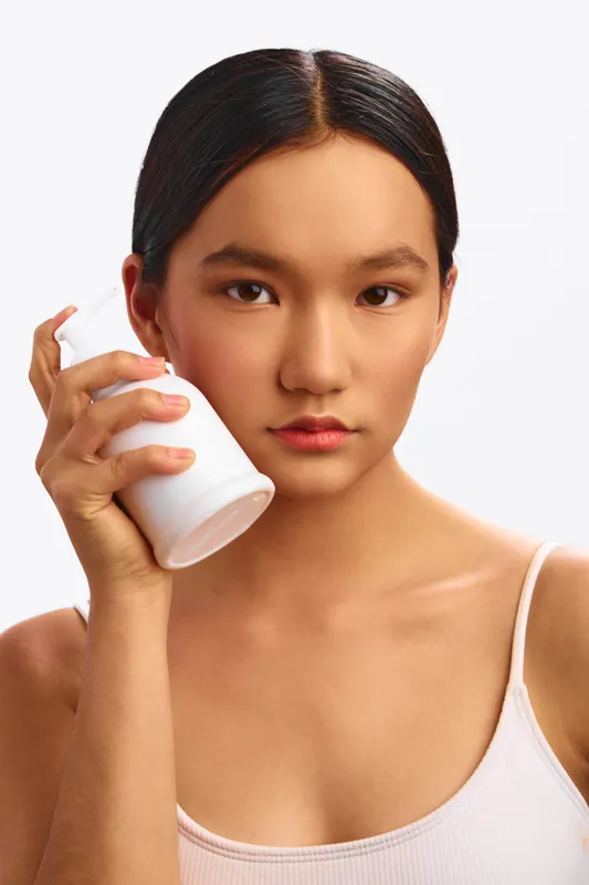Asian woman is photographed in a clean studio applying skincare product, a thoughtful expression and soft lighting convey a calm, wellness driven beauty moment.