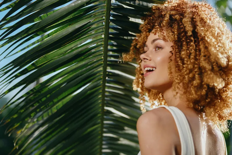 smiling woman with curly hair enjoying sunlight near green tropical palm leaves outdoors in warm summer day, natural beauty and happiness
