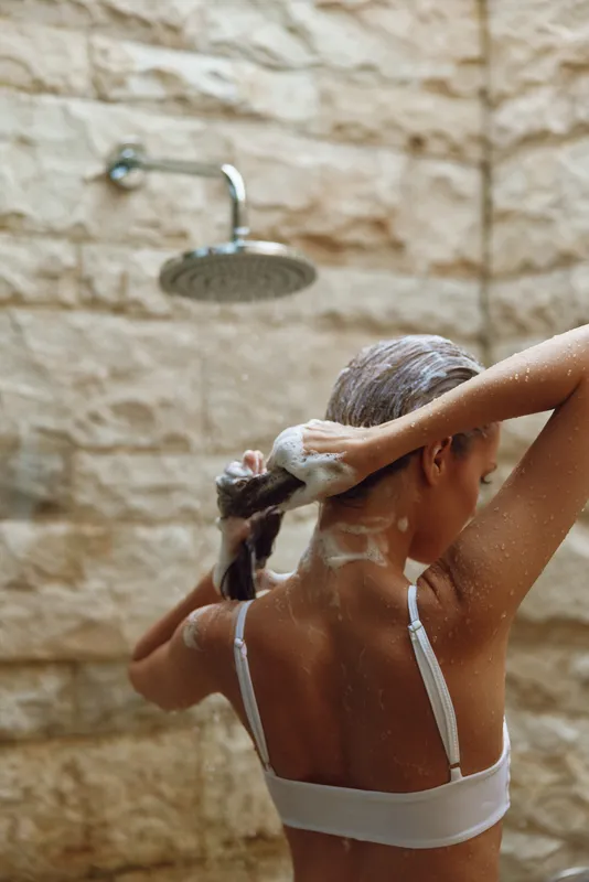 Woman washing hair in outdoor shower, natural skin care, wet hairstyle, soap suds, beige stone wall background, morning routine wellness.
