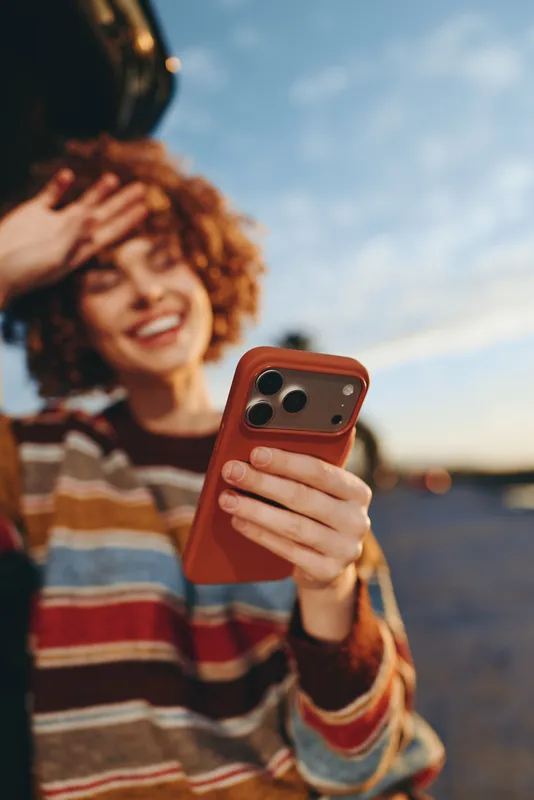 Woman lifestyle portrait outdoors, curly hair smiling while holding smartphone in her hands, candid closeup with blurred background, sunset golden light, warm leisure atmosphere