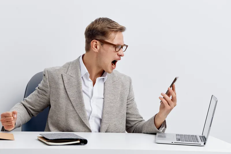 excited young man holding smartphone with laptop work desk wearing glasses gray jacket open mouth gesture cyber security concept