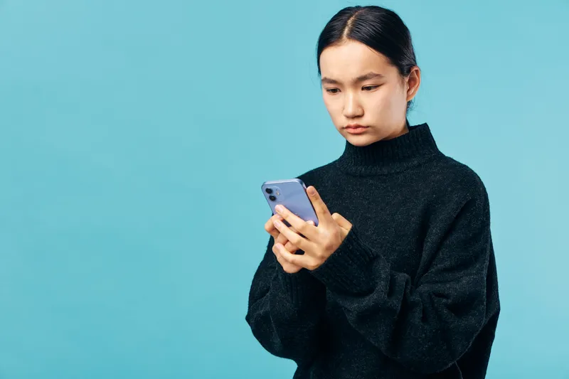 Young woman in black sweater using smartphone with focused expression on blue background. Digital communication, technology, casual style with modern device for social media or messaging.