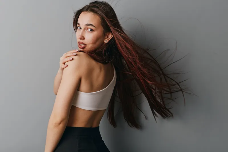 Young woman with long hair in sportswear posing with bare back and looking over shoulder against gray background for fitness and beauty concept.
