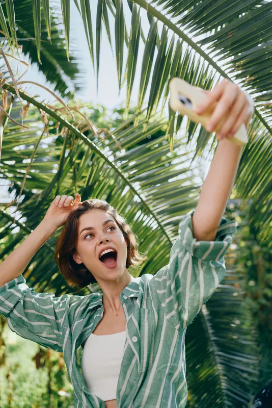 A cheerful woman takes a selfie outdoors, arms raised, capturing a sunny moment in a tropical setting