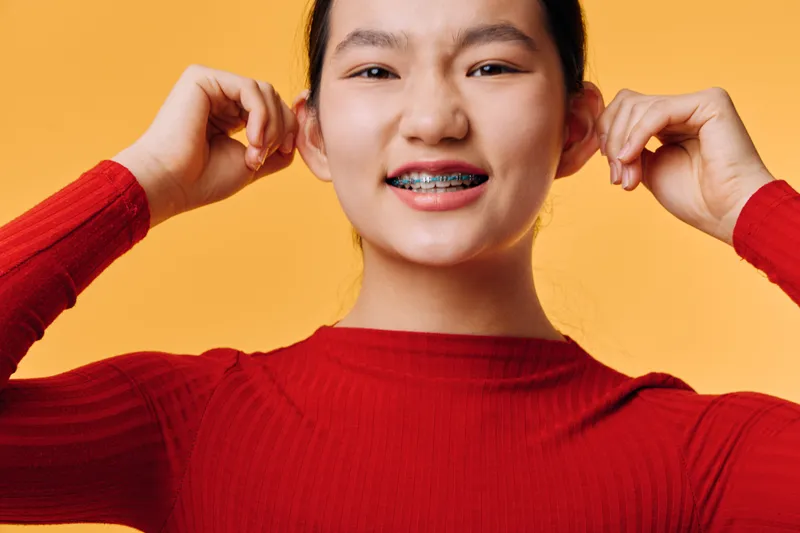Teen girl in red sweater with braces on teeth smiles and pulls ears playfully on yellow background. Portrait of adolescent person with clear skin and casual hairstyle in studio setting.