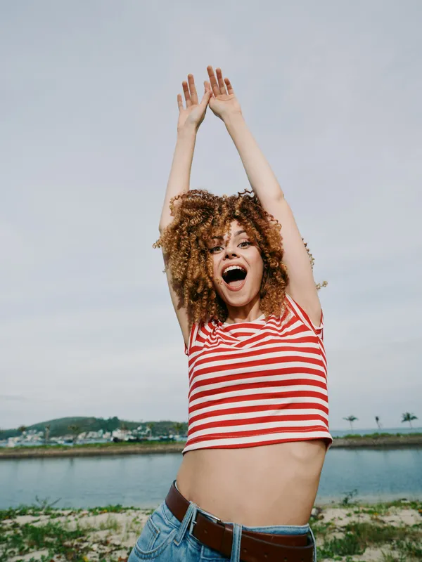 Joyful woman wearing a red striped crop top and blue jeans raises arms in celebration beside a tranquil lake under a bright sky, exuding playful summer energy and confidence.
