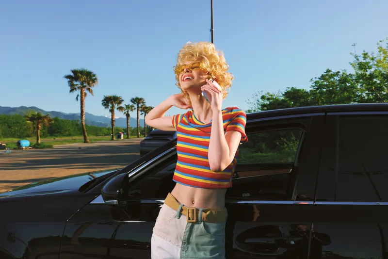 Smiling woman with curly blonde hair leaning beside a black car in a sunny outdoor scene, wearing a rainbow striped top and light shorts, conveying carefree retro summer lifestyle.