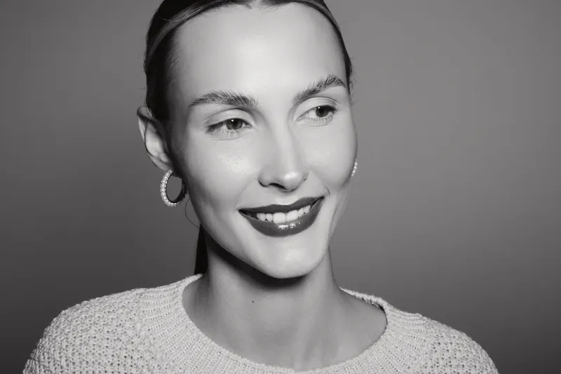 Smiling caucasian woman with fair skin and dark hair looks aside wearing hoop earrings and knitted sweater on grey background. Beauty and happiness concept.