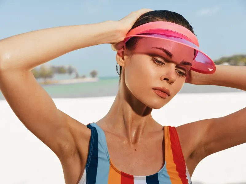 A stylish woman on a sunny beach adjusts a pink visor while wearing a bright striped swimsuit, sunlit skin, and a carefree, vibrant summer mood that conveys confidence and warmth.