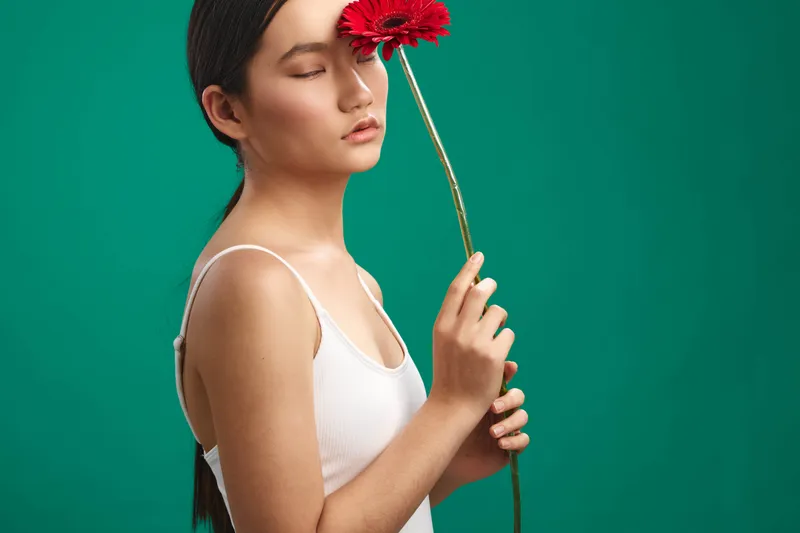 Asian woman stands in a simple studio setting, holding a red flower near her face with closed eyes, conveying calm, contemplation, and gentle elegance.