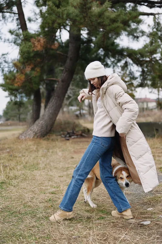 Person walking dog in a park wearing a long beige coat and beanie, jeans and boots, enjoying a casual outdoor moment among trees and open autumn scenery