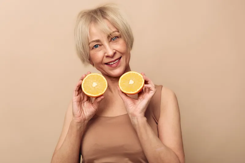 Happy mature woman with short blonde hair holds two orange halves near face smiling gently on beige background. Concept of healthy lifestyle, vitamin C, nutrition, and wellness for older adults.