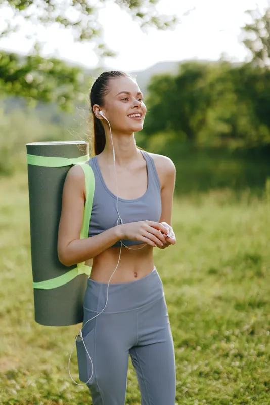 Young athletic woman smiling outdoors with yoga mat, dressed in stylish fitness attire, representing wellness and active lifestyle in serene environment
