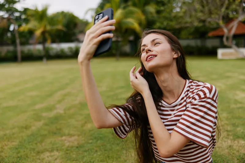 Женщина в футболке, природа, улыбка, пальма, просматривает телефон на открытом воздухе, джинсы, парк, прогулка, зелень, блогер, образ жизни, сотовый телефон, лето, трава, на улице, дерево…