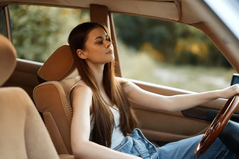 Young woman with long hair sitting in a car, wearing casual attire, exuding a calm and confident expression in a soft, natural environment.
