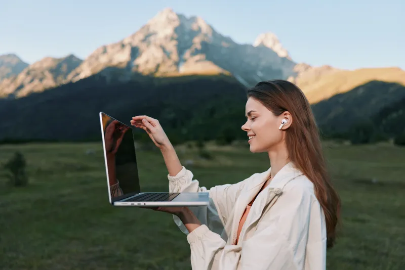 A focused professional woman stands outdoors, holding a laptop and reaching toward the screen, smiling softly in a bright scenic setting.
