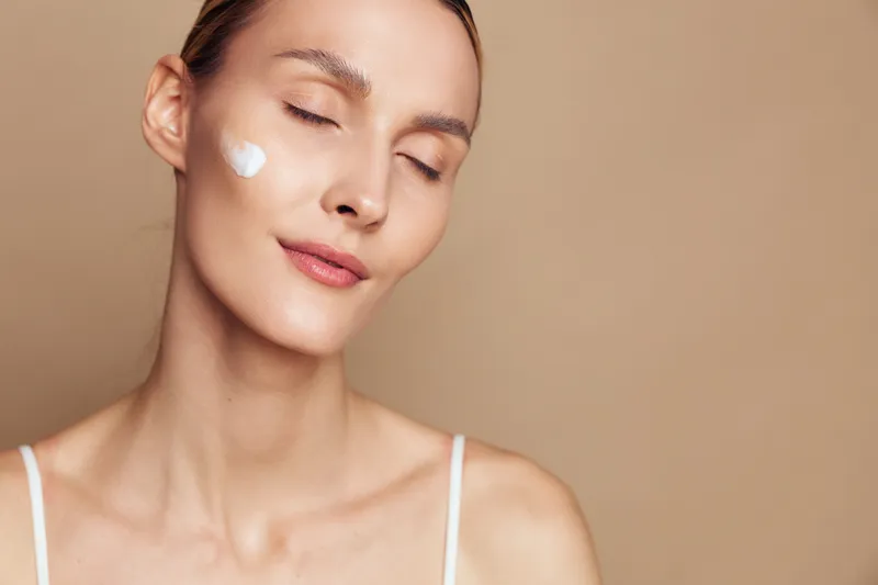 Close up of a young adult woman with a dab of cream on her cheek, serene expression, soft beige background, minimal makeup, skincare moment, gentle self care.
