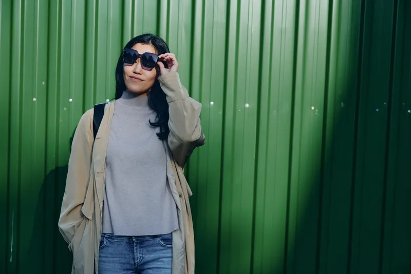Woman in sunglasses and beige trench coat stands beside a green wall, showcasing casual street style, chic layered outfit, and confident pose, urban vibe and sunny ambiance for fashion editorial