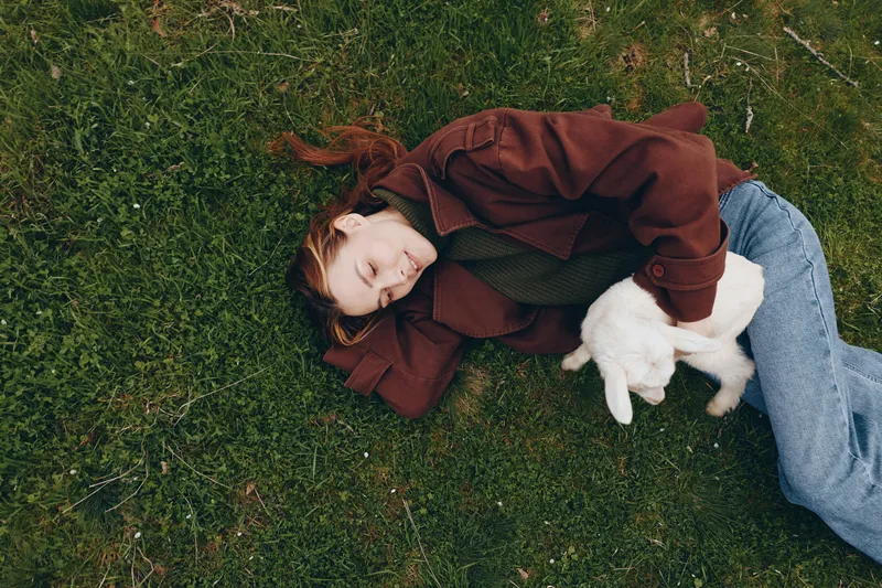 A girl lying on green grass outdoors with a goat kid by her side, a calm lifestyle moment showing companionship, gentle affection, and simple wholesome outdoor life.