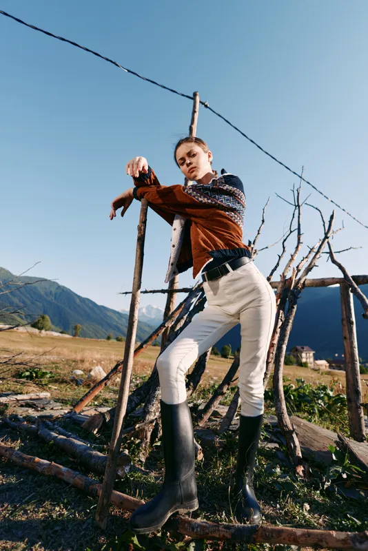 Woman standing by wooden fence in countryside wearing sweater, white pants and boots, posing outdoors near mountain under clear blue sky for rustic fashion portrait and lifestyle
