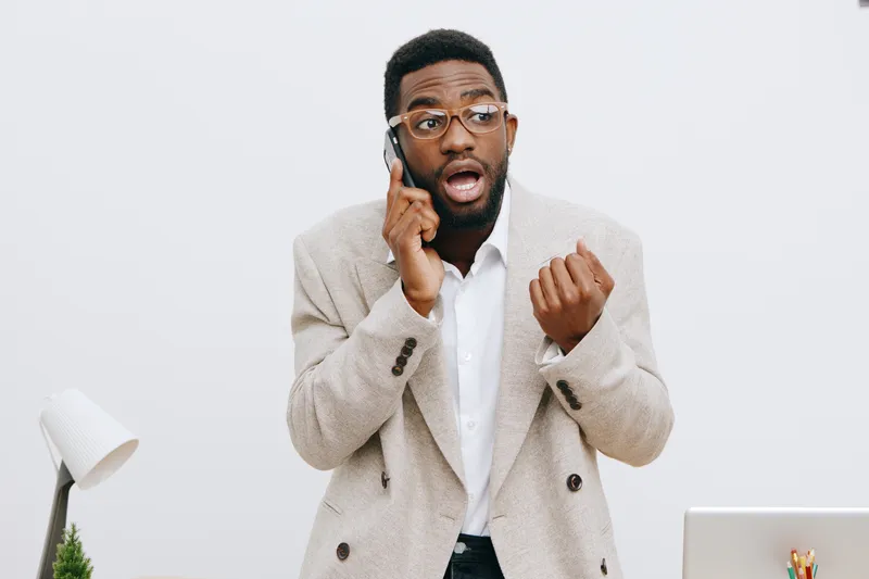 Frustrated young man with glasses and beard talking on mobile phone while gesturing with hand in modern office environment, wearing beige blazer and white shirt. People conflict situation concept