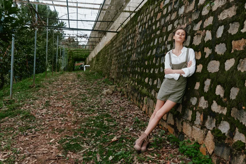 Stylish female model wearing a casual outfit poses against a textured stone wall in a lush outdoor setting, exuding confidence and serenity, perfect for fashion concepts
