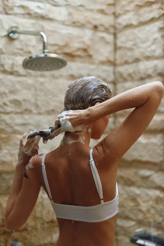 woman washing hair in shower with shampoo foam, natural stone background, wet skin and relaxed posture in warm light