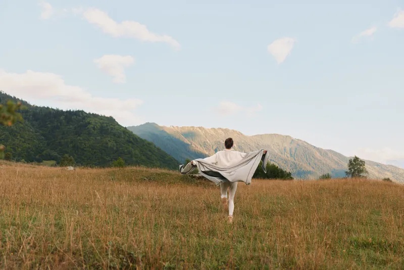 Young woman stands in a sunlit field with arms outstretched, a flowing shawl catching the breeze, while distant mountains frame a peaceful, expansive countryside scene.
