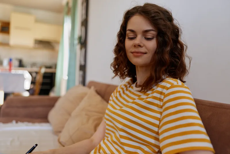 Woman at home in a casual striped shirt sits on a sofa and writes in a notebook with a pen, capturing a relaxed lifestyle moment in warm natural light indoors.