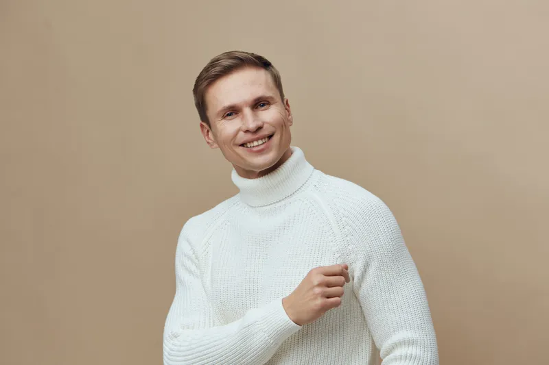 Smiling young man wearing cozy white turtleneck sweater against beige background, looking at camera, cheerful expression, casual style, portrait photography, lifestyle shot, studio lighting