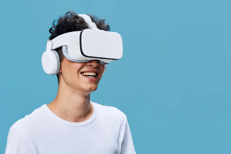 Smiling man wearing virtual reality headset and headphones enjoys immersive digital experience against blue background with casual white shirt, modern technology concept.