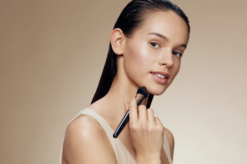 Young woman applying makeup with a brush, showcasing a natural beauty look on a neutral background, ideal for beauty and skincare industries