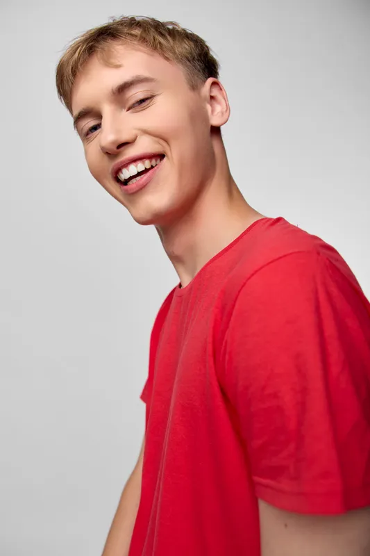 Smiling young man in a red shirt stands in a bright studio, showcasing casual fashion, happiness, and a confident, approachable vibe with a clean modern look for lifestyle stock