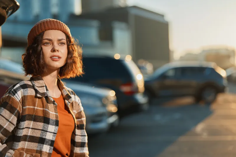 woman portrait candid authenticity golden urban lifestyle, young woman in beanie and plaid jacket at golden hour in a parking lot, warm glow, thoughtful candid expression, emotional storytelling