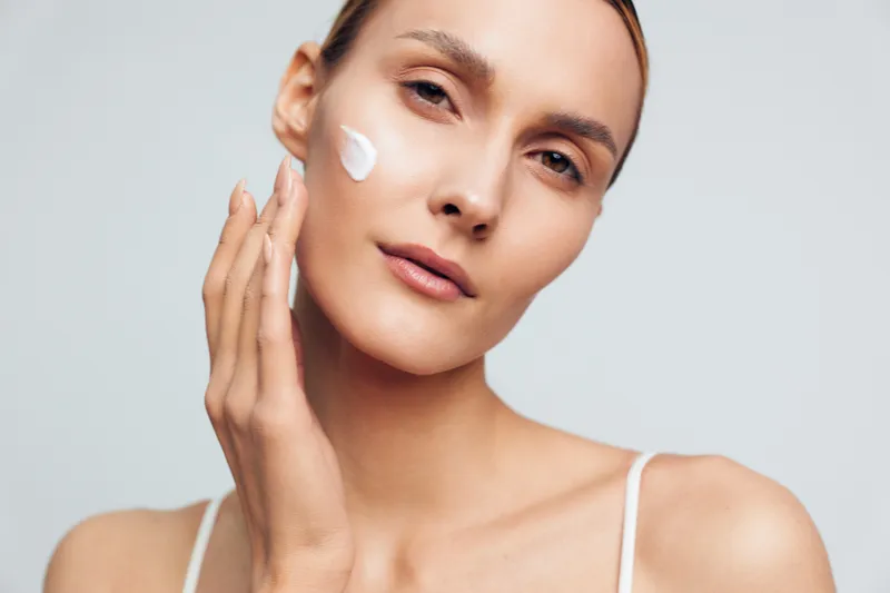 A young woman applies moisturizing cream to her cheek with her hand against a plain light background. The concept focuses on skincare, beauty routine, and healthy skin care.