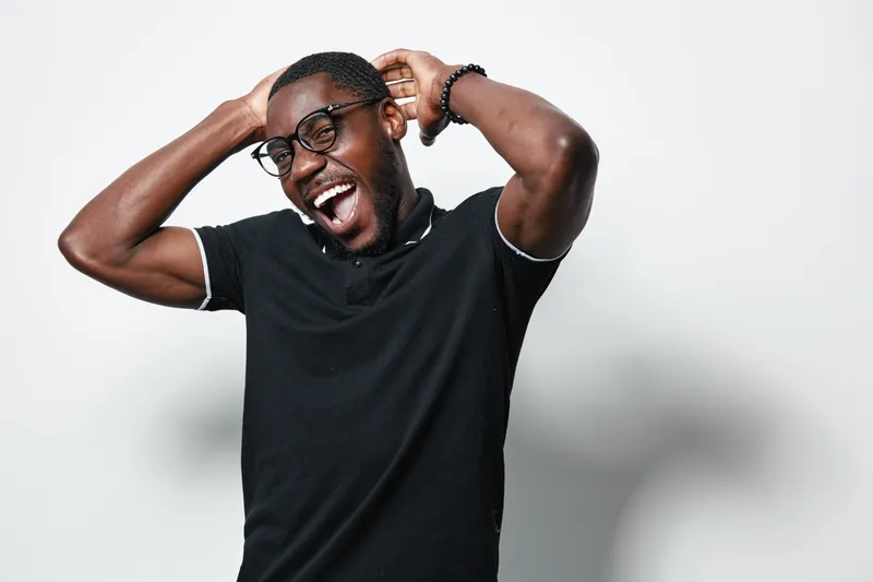 Young black man with dark skin and glasses laughing with hands behind head wearing black shirt on white background. Happy African ethnicity male expressing joy and relaxation.
