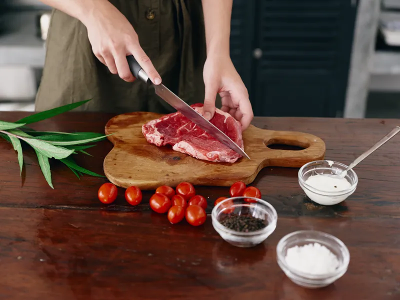 Женщина с ножом в руке нарезает стейк из мяса для жарки на кухне
