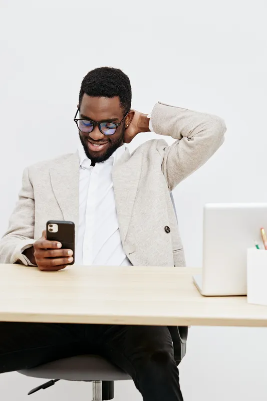 Smiling man using smartphone at desk in modern office, dressed in smart casual blazer and white shirt, sitting in front of laptop, looking pleased with message or notification, professional workplace 