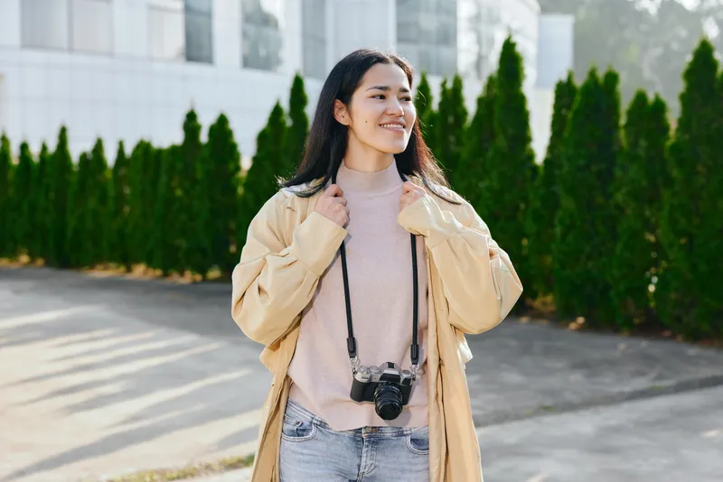 Smiling woman wearing a beige coat and soft pink sweater, with a camera hanging from her neck, stands outdoors beside tall evergreens, ready for a casual fashion shoot and street photography in
