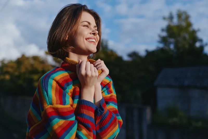 Woman, smiling woman in a rainbow striped sweater with short brown hair, outdoor portrait of a young adult holding collar, joyful expression, casual lifestyle and fashion portrait.