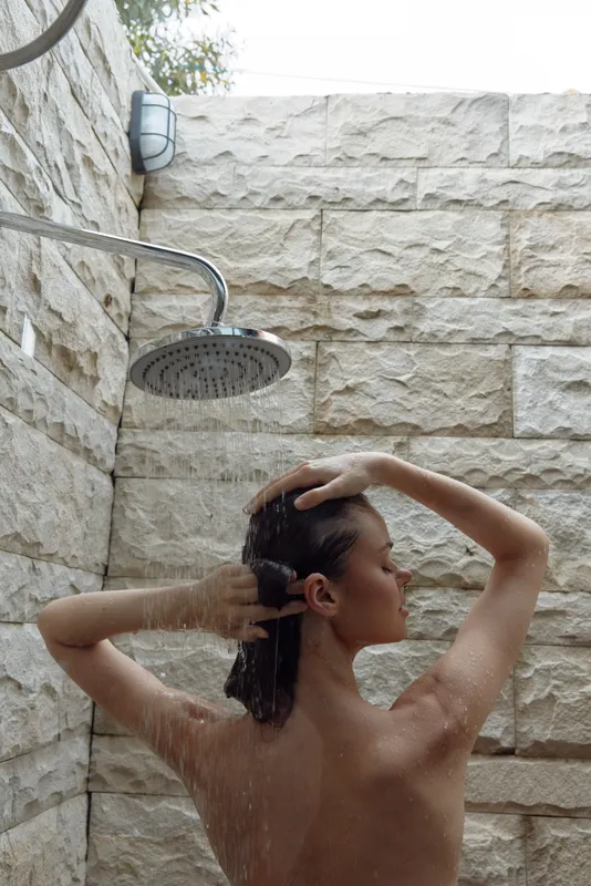 woman washing hair under outdoor shower stone wall textured surface water droplets natural light wellness and relaxation