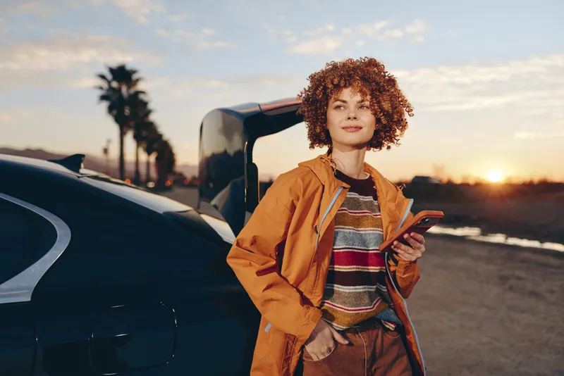 Woman with curly hair wearing jacket near car, smiling in lifestyle travel scene at sunset along road, she holds phone and keys by the open door in outdoor evening light with warm golden sky