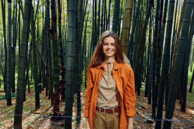 woman bamboo forest smile portrait outdoors nature travel: Young woman smiling in a dense bamboo grove wearing an orange jacket, casual outfit, standing on leaf covered forest floor for outdoor