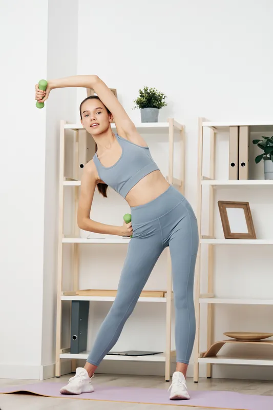 Fitness woman, home workout, dumbbell exercise, athletic pose in a bright space. Woman in light blue athletic wear stretches with a dumbbell beside a yoga mat and clean minimalist shelves.