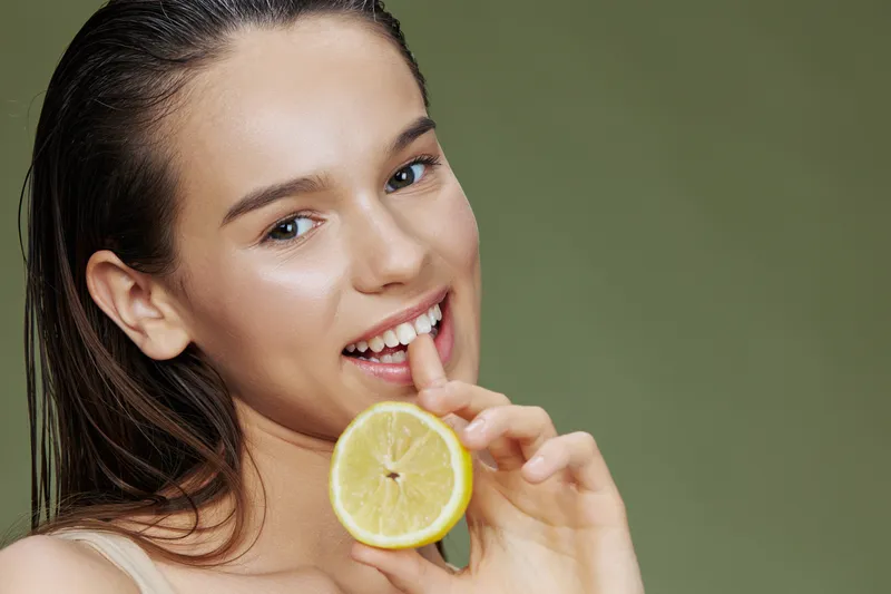 Брюнетка держит лимон рядом с лицом, чистая кожа, уход за здоровьем, зелёный фон